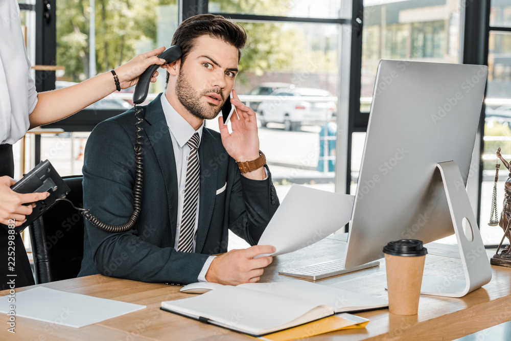 shocked businessman talking by smartphone and secretary holding handset of stationary telephone near his ear shocked businessman talking by smartphone and secretary holding handset of stationary telephone near his ear