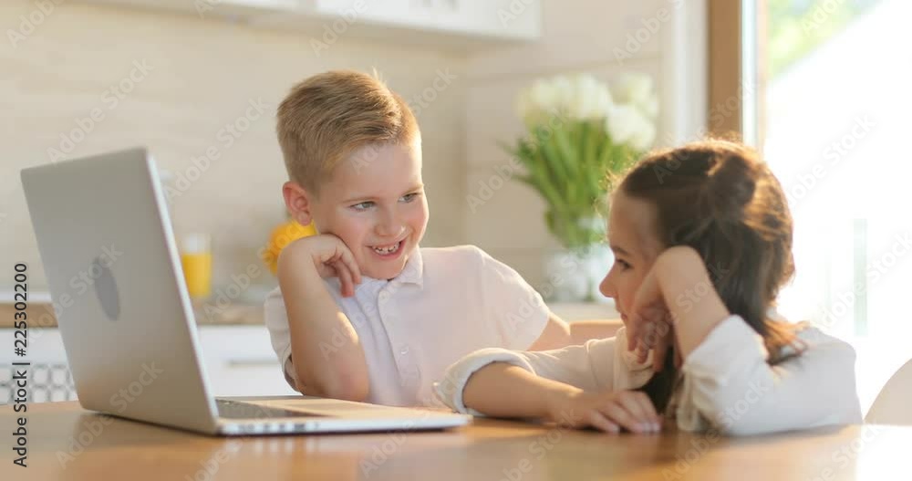 Portrait of two smiling school kids using laptop computer in kitchen. Home, school, education concept. Boy and girl operating laptop Portrait of two smiling school kids using laptop computer in kitchen. Home, school, education concept. Boy and girl operating laptop