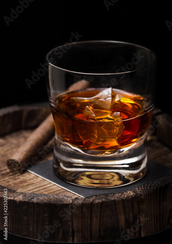 Glass of whiskey with ice cubes and cigar on top of wooden barrel. Cognac brandy drink