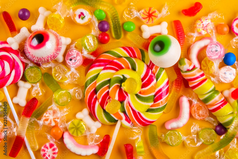 Assorted teeth and eyeball shaped candy spread on yellow background, jelly spider, gummy worms, sugar bones, round lollipop and other mixed candy. Top view, copy space, close up, flat lay.