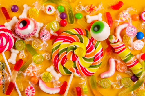 Assorted teeth and eyeball shaped candy spread on yellow background  jelly spider  gummy worms  sugar bones  round lollipop and other mixed candy. Top view  copy space  close up  flat lay.