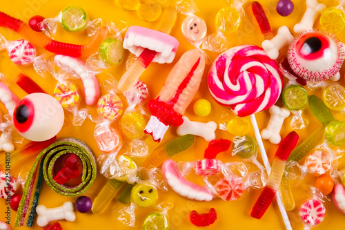 Assorted teeth & eyeball shaped candy spread on yellow background, jelly spider, gummy worms, sugar bones, round lollipop and other mixed candy, bloody finger. Top view, copy space, close up, flat lay