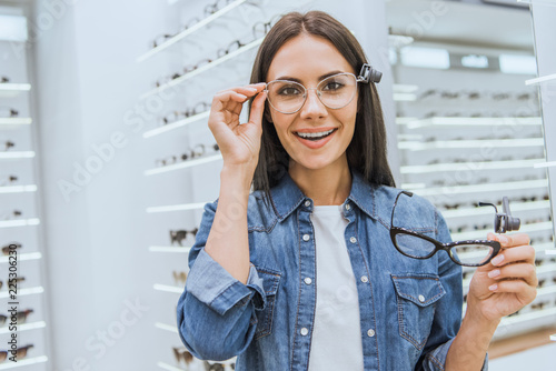 Photography cheerful young woman choosing eyeglasses and looking at camera in ophthalmic sho