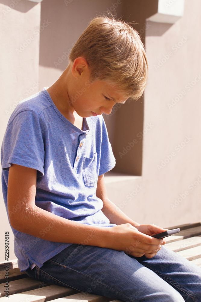 Boy with smartphone sitting on stairs outdoor. Kid watching to screen, reading, typing, playing games
