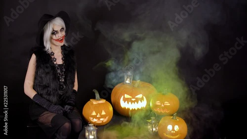 Scary beautiful girl witch celebrates halloween with funny glowing burning pumpkins in smoke. Female sorceress with gray hair scares, conjures, casts a spell