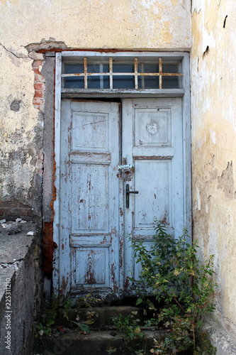 Light blue old dilapidated wooden doors locked with small padlock with windows above secured with rusted metal bars on abandoned house with stone steps partially covered with plants in front © hecos