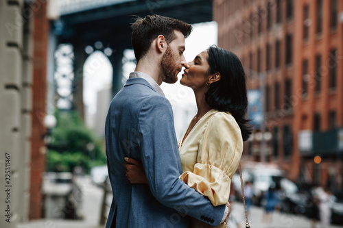 Love story in New York. Gorgeous couple of American man with beard and tender Eastern woman hug each other before the cityscape of Brooklyn bridge somewhere in New York © IVASHstudio