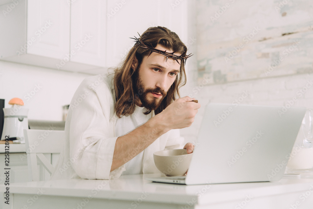 focused Jesus eating corn flakes on breakfast at table with laptop in kitchen at home focused Jesus eating corn flakes on breakfast at table with laptop in kitchen at home