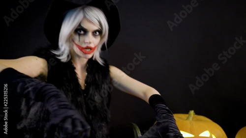 Female sorceress with gray hair scares, conjures, casts a spell. Scary beautiful girl witch celebrates halloween with funny glowing burning pumpkins in smoke. Gates of hell in fire