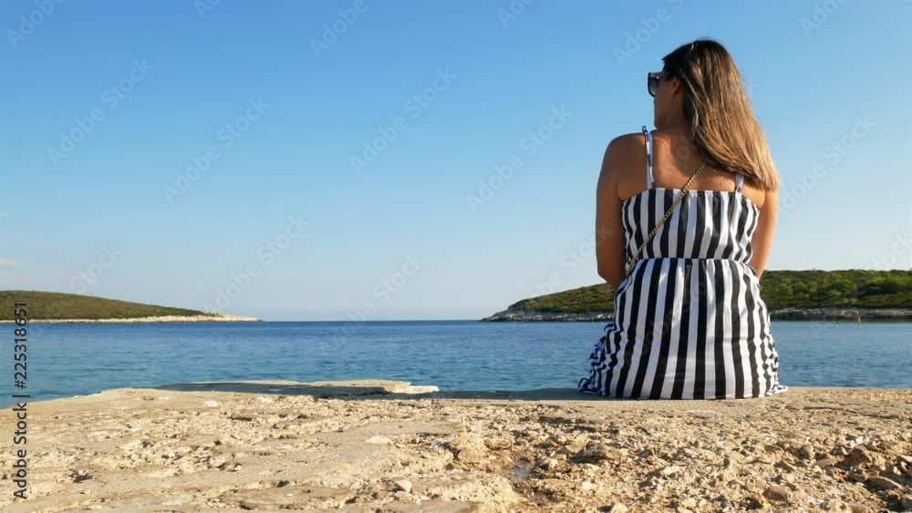 Young caucasian woman sitting alone on stone dock on Croatian coast and looking at blue sea Young caucasian woman sitting alone on stone dock on Croatian coast and looking at blue sea