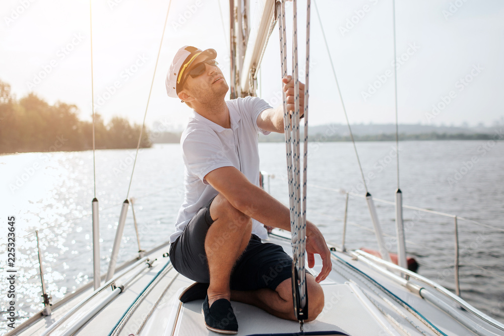 Sailor sits on board of yacht and look up. He holds ropes in hands. Guy is concentrated. It is beautiful and sunny day outside.