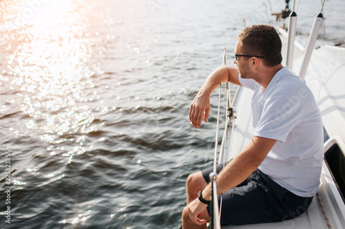 Young man sits at adge of yacht board and looks forward on sun. He leans on railing. Guy is calm and peceful. He is chilling. Water is calm. photo
