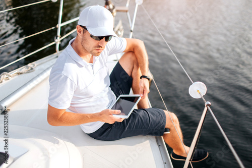 Picture of young man sits at edge of yacht board and hold tablet. It has dark screen. Man wears sunglasses and white cap with shirt. He is serious and confident. Sailor get some rest.