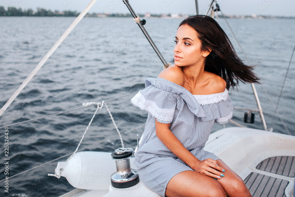 Amazing and beautiful young woman sitting on board of yacht and look to left. She is calm and peaceful. Woman is alone.