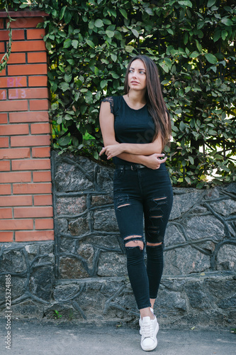 A brunette female with long hair wearing a blank black t-shirt i while standing on a concrete wall background on a street. Empty space for text or design.