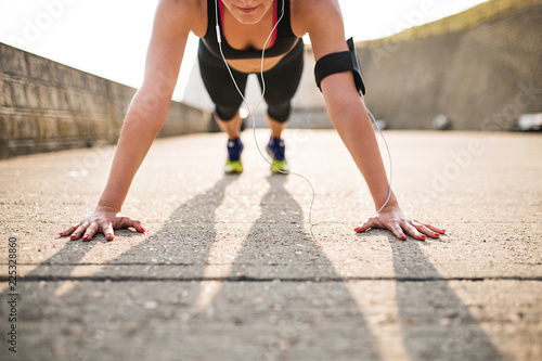 Young sporty woman runner with smartphone doing push-ups outside.