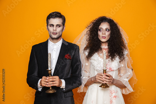 Photo of attractive zombie couple bridegroom and bride wearing outfit and halloween makeup holding candles, isolated over yellow background photo