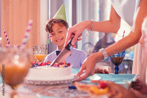 Cutting cake. Cheerful satisfied boy smiling while looking at his mother slicing his big birthday cake