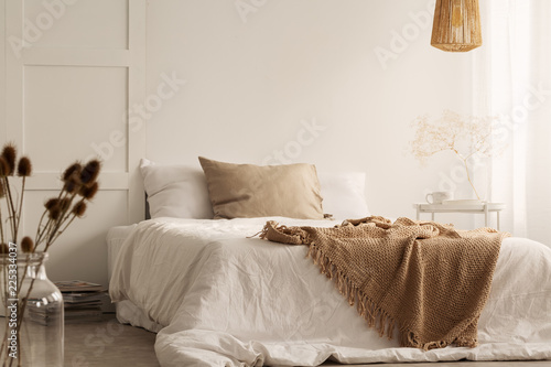 Flowers and lamp in white natural bedroom interior with blanket and pillows on bed. Real photo