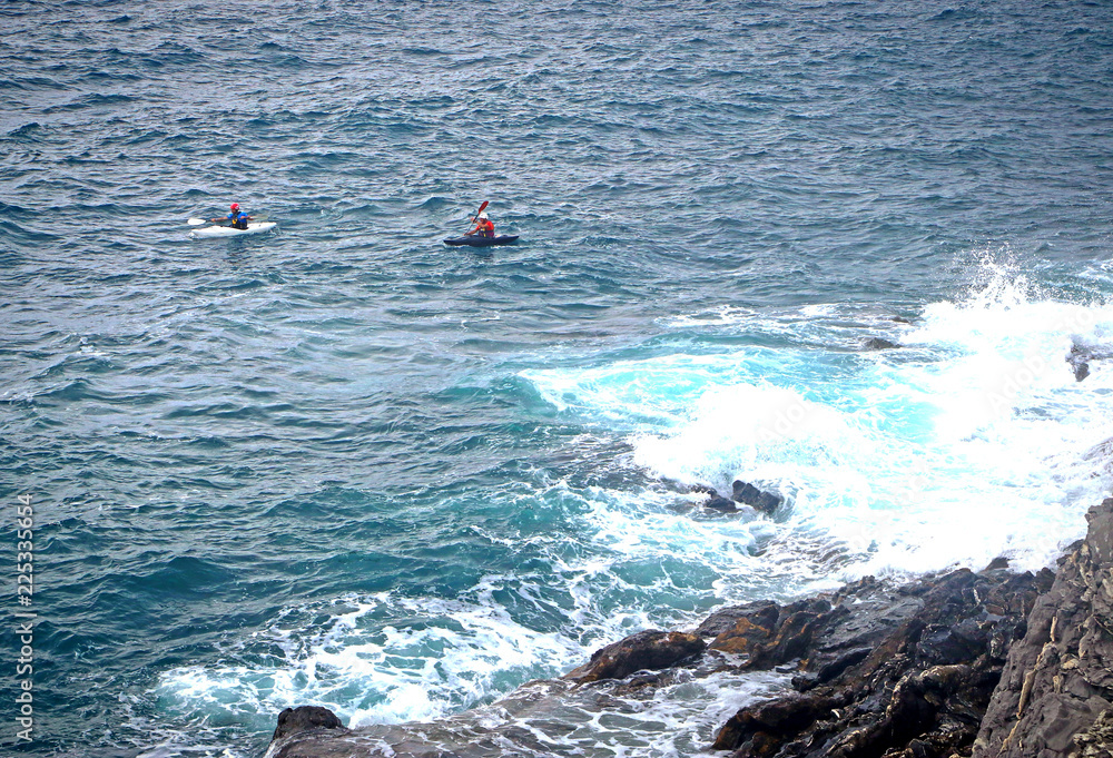 two people in kayak paddle on the stormy sea with breaking waves along the rocky coast