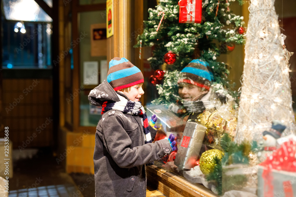 Funny happy child in fashion winter clothes making window shopping decorated with gifts, xmas tree