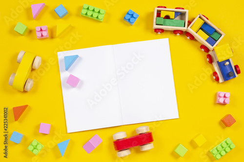Early education background. Wooden train, car, colorful bricks and blocks with open blank notebook on yellow background