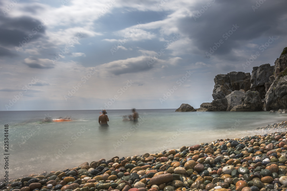 People at Mediterranean beach. Slow motion (speed) long exposure. Colorful pebbles and silky waves of blue sea on the shore. Texture, background concept.