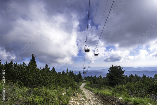 Onboard a ski lift going up a mountain at summertime with people. Cloudy and forest in Rila mountain in Bulgaria - the way to the seven Rila lakes. Lifting