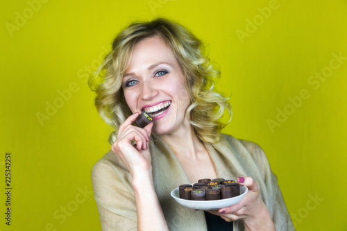 A woman with a beautiful smile is eating candy. A beautiful girl with healthy teeth bites chocolate and holds a plate of sweets on a yellow background.