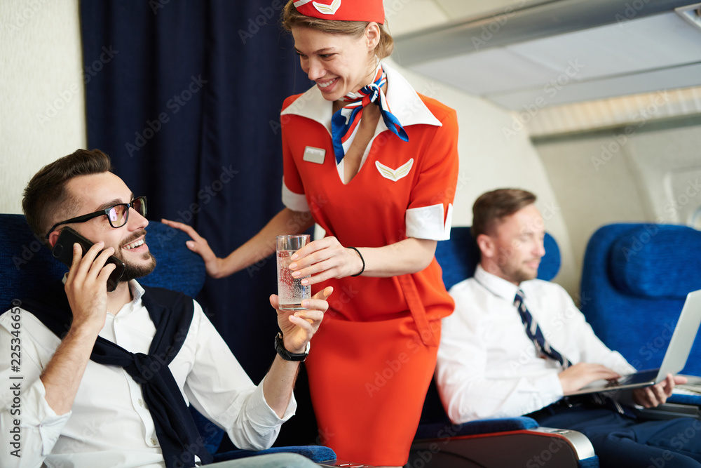 Portrait of smiling flight attendant serving glass sparkling water to handsome businessman enjoying first class trip, copy space - obrazy, fototapety, plakaty 