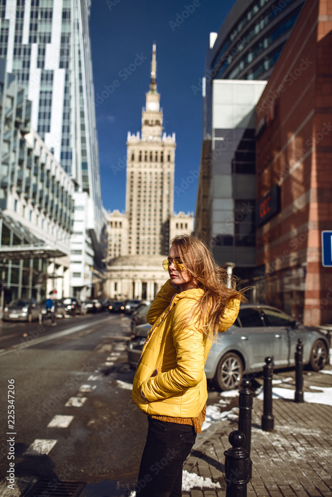 A young European woman, traveling, with long blond hair, wearing a yellow jacket, yellow sunglasses walking down the city center street, street shooting. Even light.