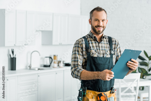 smiling handsome plumber holding clipboard and looking at camera in kitchen photo