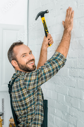 Fototapeta smiling handsome repairman hammering nail in white wall and looking at camera