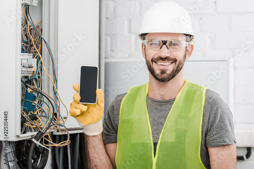 smiling handsome electrician showing smartphone with blank screen near electrical box in corridor