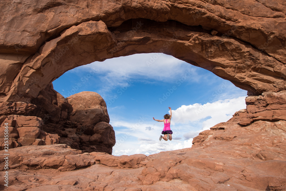 Adult woman jumps inside of Skyline Arch in Arches National Park, in Utah. Beautiful blue sky peeking from behind the arch