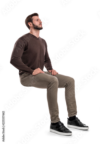 An isolated bearded man in casual wear sits on a white background with hands on his thighs and looks up. photo
