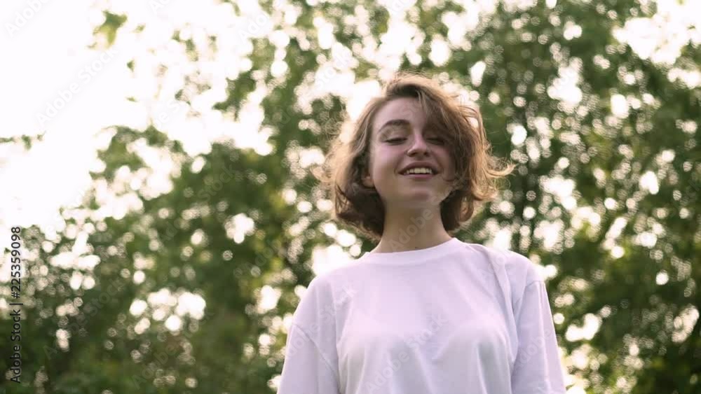Cute young girl in white t shirt walking in summer park, playing with her short fair hair and putting on pink sunglasses. Concept of fashion and happiness. Tracking slow motion low angle shot Cute young girl in white t shirt walking in summer park, playing with her short fair hair and putting on pink sunglasses. Concept of fashion and happiness. Tracking slow motion low angle shot