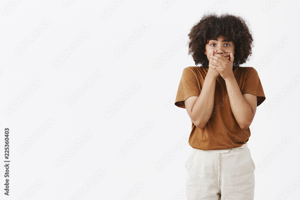 Portrait of shocked speechless African American woman witnessing shocking scene covering mouth with both hands standing in stupor over gray background and looking at camera stunned and shook Portrait of shocked speechless African American woman witnessing shocking scene covering mouth with both hands standing in stupor over gray background and looking at camera stunned and shook
