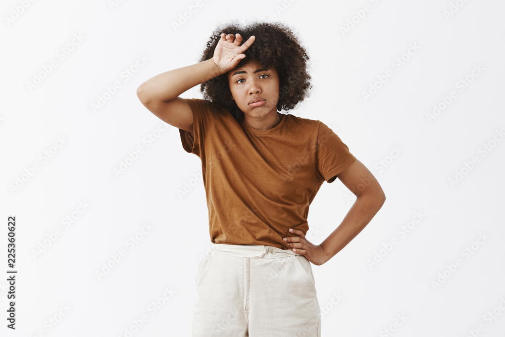 Tired stylish and cute dark-skinned female model with curly hairstyle whiping sweat from forehead breathing out, pouting and holding hand on waist being exhausted from carrying heavy things Tired stylish and cute dark-skinned female model with curly hairstyle whiping sweat from forehead breathing out, pouting and holding hand on waist being exhausted from carrying heavy things