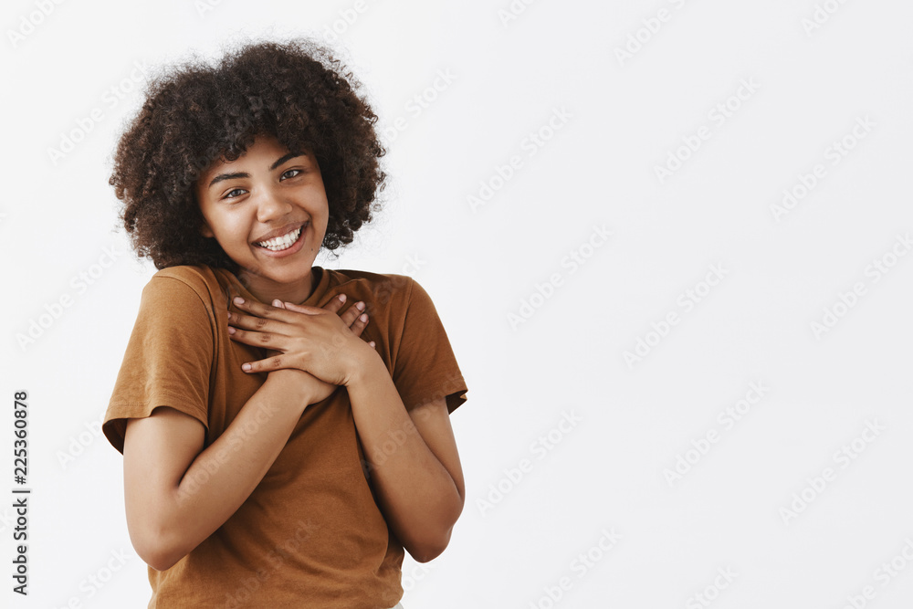 Cute and tender romantic dark-skinned female student with afro hairstyle in brown t-shirt holding hands on heart tilting head flirty and smiling being touched and pleased over gray wall Cute and tender romantic dark-skinned female student with afro hairstyle in brown t-shirt holding hands on heart tilting head flirty and smiling being touched and pleased over gray wall