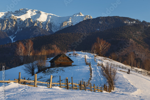 Alpine scenery with rustic wooden cottage and snow capped ridge of the Bucegi mountains in the sunset light in a cold winter evening, in Bran, Romania.