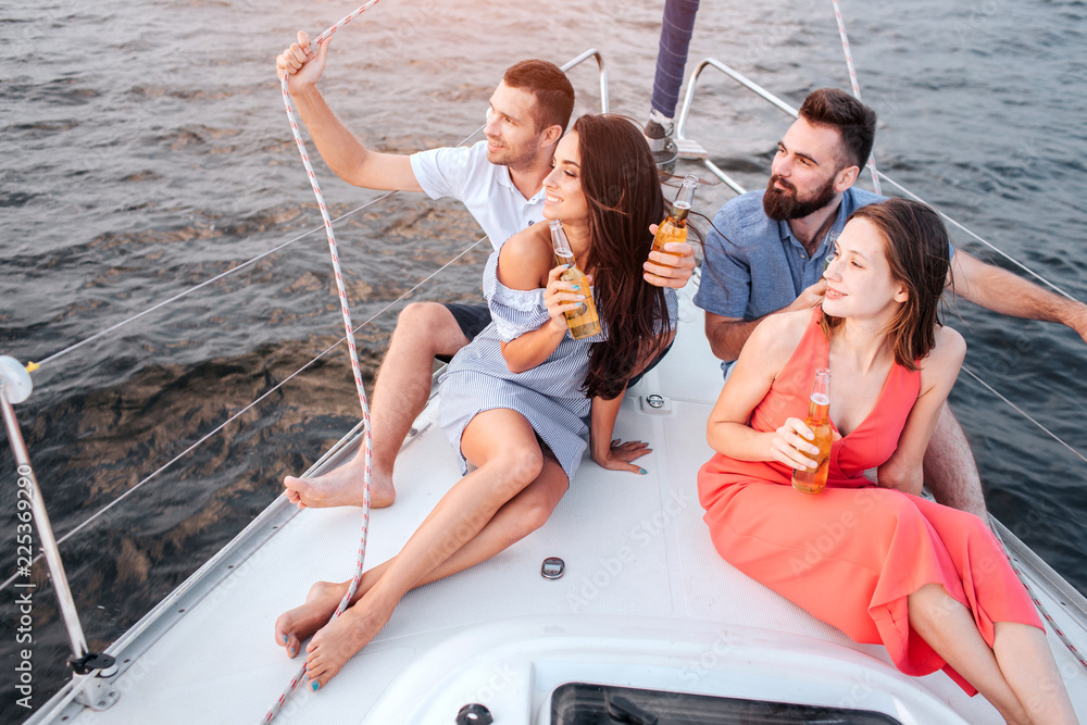 Two couples are sitting on bow of yacht. They look to left. Women are excired. Men are happy too. They watch on sunset.