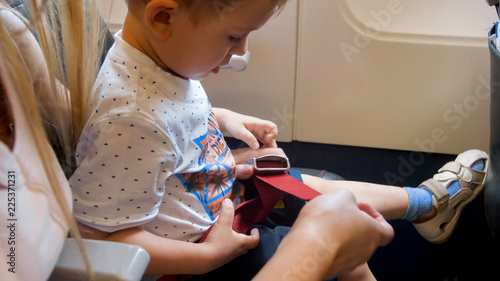 Young mother fastening and pulling safety seat belt of her child in aircraft before taking off photo