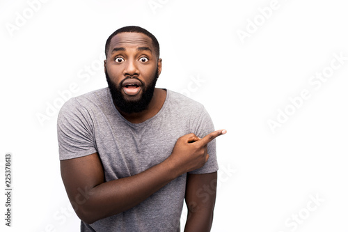 Headshot of attractive African man looking at camera with amazed expression eyes and full disbelief, pointing his index finger at blank copy space wall, shocked with content