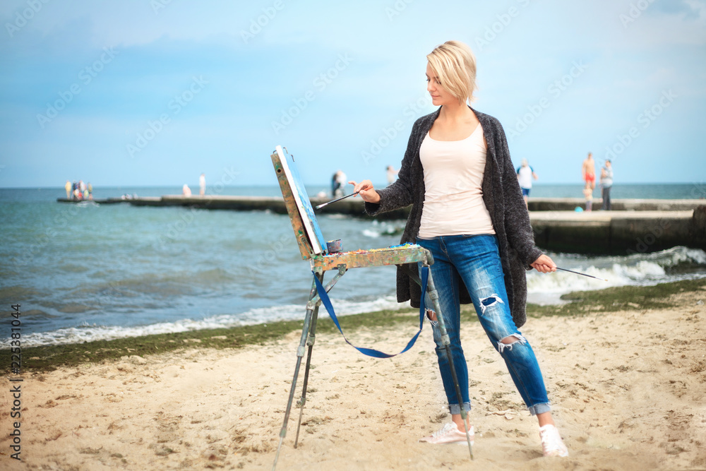 Beautiful young woman painting on the background of the sea and sky. The concept of creativity.