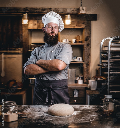 Professional baker in cook uniform posing with crossed arms near table with ready dough in the bakery.