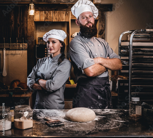 Chef with his assistant in cook uniform posing with crossed arms near table with ready dough in the bakery. photo