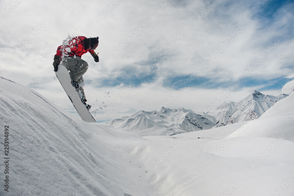 Jumping snowboarder keeping his hand on the snowboard on the background of mountains and sky