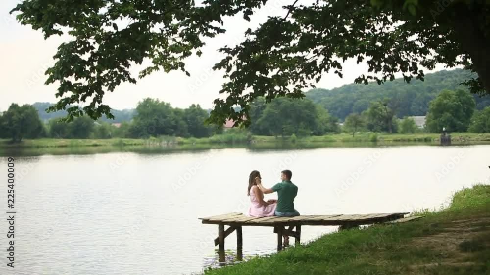 Beautiful youn couple sit on the pier near the lake. Romantic atmosphere. Back biew Beautiful youn couple sit on the pier near the lake. Romantic atmosphere. Back biew