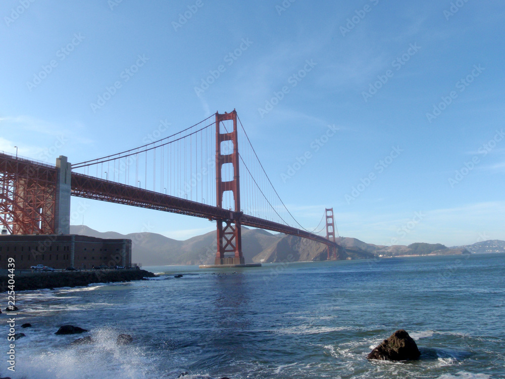Naklejka premium Wave breaks on shore with Golden Gate Bridge, Marin County and Fort Point in distance